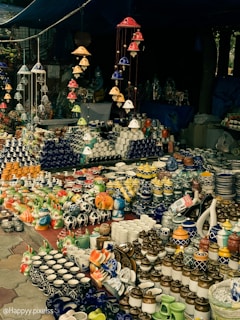 A market stall displaying an array of vibrant ceramic items, including mugs, pots, and decorative figurines. The arrangement includes various colors and patterns, with items neatly organized on tables. Overhead, colorful ceramic wind chimes hang, adding to the overall aesthetic.