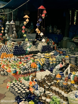 A market stall displaying an array of vibrant ceramic items, including mugs, pots, and decorative figurines. The arrangement includes various colors and patterns, with items neatly organized on tables. Overhead, colorful ceramic wind chimes hang, adding to the overall aesthetic.
