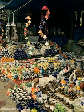 A market stall displaying an array of vibrant ceramic items, including mugs, pots, and decorative figurines. The arrangement includes various colors and patterns, with items neatly organized on tables. Overhead, colorful ceramic wind chimes hang, adding to the overall aesthetic.