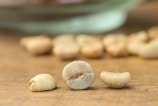 Three unroasted coffee beans are placed on a wooden surface, with more scattered and blurred into the background. The beans have a greenish hue and display their natural imperfections.
