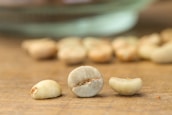 Three unroasted coffee beans are placed on a wooden surface, with more scattered and blurred into the background. The beans have a greenish hue and display their natural imperfections.