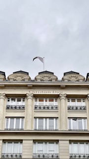 A grand architectural facade with classical design elements features columns and ornate decorations. The upper part displays the name Christian Dior, with decorative stonework aligned above windows and a flag waving on top.