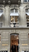 The image depicts the exterior of a high-end store with the brand name 'GUCCI' prominently displayed. The architecture features ornate designs with arched windows and detailed stonework. There are two visible floors with the ground floor window showing a well-lit interior and a group of people looking at the displays. The upper floor has a window with a 'GUCCI' awning.