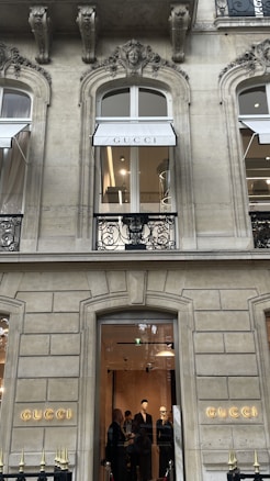The image depicts the exterior of a high-end store with the brand name 'GUCCI' prominently displayed. The architecture features ornate designs with arched windows and detailed stonework. There are two visible floors with the ground floor window showing a well-lit interior and a group of people looking at the displays. The upper floor has a window with a 'GUCCI' awning.