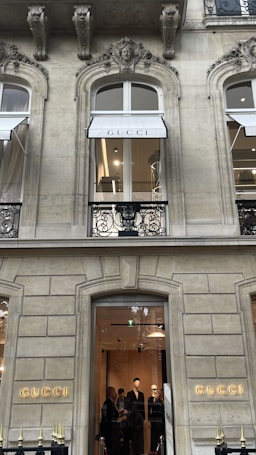 The image depicts the exterior of a high-end store with the brand name 'GUCCI' prominently displayed. The architecture features ornate designs with arched windows and detailed stonework. There are two visible floors with the ground floor window showing a well-lit interior and a group of people looking at the displays. The upper floor has a window with a 'GUCCI' awning.