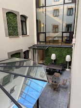 An interior courtyard featuring vertical gardens growing on the walls, large glass windows showcasing reflections, and a seating area with tables and chairs. Two tall, sculptural floor lamps enhance the modern design. The floor is tiled, adding a geometric pattern to the overall clean aesthetics.