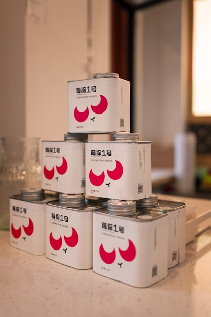 A stack of small metal cans with a clean, modern design featuring bright red abstract shapes and black typography. Each can has a silver screw-on lid and is neatly arranged on a countertop, suggesting an organized setting.