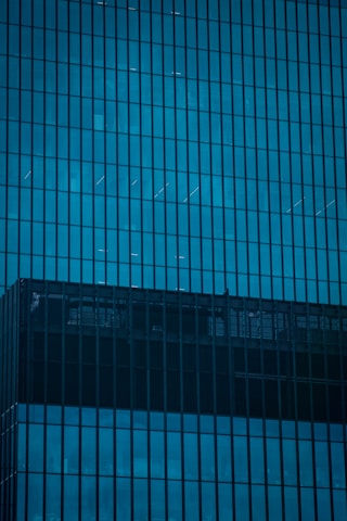 A grid of blue-tinted glass windows forming the façade of a modern high-rise building. The reflective surfaces create patterns with lines and shadows, highlighting architectural details.