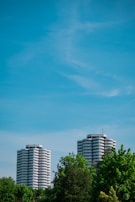 Two tall, cylindrical apartment buildings rise above lush green trees under a clear blue sky. The buildings feature multiple balconies with a modern design.