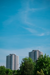 Two tall, cylindrical apartment buildings rise above lush green trees under a clear blue sky. The buildings feature multiple balconies with a modern design.