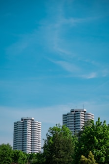 Two tall, cylindrical apartment buildings rise above lush green trees under a clear blue sky. The buildings feature multiple balconies with a modern design.