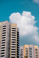 Tall residential apartment buildings with a modern architectural style are set against a backdrop of a bright blue sky with large, fluffy clouds. The buildings feature multiple balconies and a combination of beige, gray, and light yellow colors.