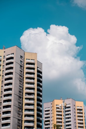 Tall residential apartment buildings with a modern architectural style are set against a backdrop of a bright blue sky with large, fluffy clouds. The buildings feature multiple balconies and a combination of beige, gray, and light yellow colors.