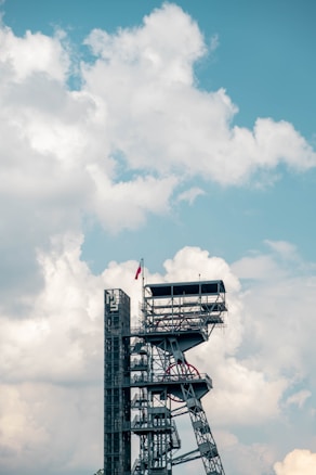 A tall, industrial-looking structure with multiple platforms and staircases extends against a backdrop of large, fluffy white clouds in a light blue sky. A red flag is positioned at the top of the structure.