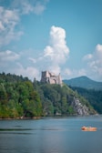 A serene landscape featuring a historic castle situated on a lush, green hill beside a large body of water. The sky is bright with scattered clouds, and a small orange boat carrying people is visible on the water.