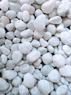 A collection of white pebbles is closely packed together. Each pebble varies slightly in size and shape, creating a textured surface. The pebbles have smooth surfaces and a consistent light color, with subtle variations in shade due to natural imperfections.