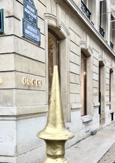 A view of a luxury store facade on Avenue Montaigne, featuring elegant stone architecture with large windows. A metal fence with a golden pointed finial is visible in the foreground.
