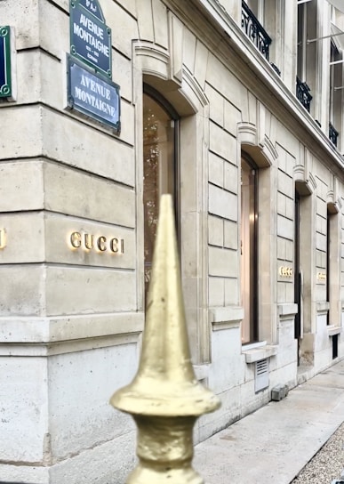 A view of a luxury store facade on Avenue Montaigne, featuring elegant stone architecture with large windows. A metal fence with a golden pointed finial is visible in the foreground.