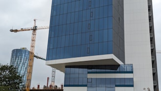 A modern high-rise building with a glass facade is prominently featured, alongside a large construction crane. In the background, another building with a similar glass appearance is visible, and there are trees partially visible at the bottom.