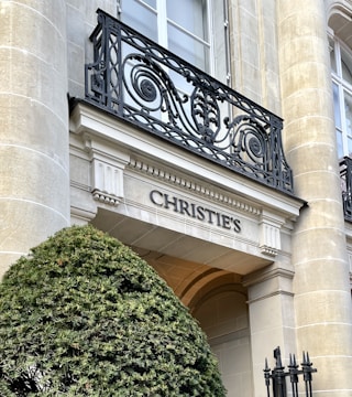 A stone facade with ornate black wrought iron railings and the name 'CHRISTIE'S' engraved. In front, there is a neatly trimmed shrub and portions of classical architectural elements, including columns with detailed capitals.