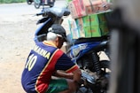 A man wearing a jersey with the name 'Kumarna' and the number 10 is crouched down next to a blue motorbike loaded with stacked boxes, including brands like Le Mintea. The motorbike is parked on a dirt surface with a road visible in the background.