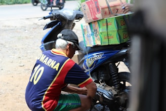 A man wearing a jersey with the name 'Kumarna' and the number 10 is crouched down next to a blue motorbike loaded with stacked boxes, including brands like Le Mintea. The motorbike is parked on a dirt surface with a road visible in the background.