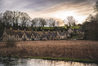 A picturesque row of rustic stone cottages with thatched roofs situated in a tranquil rural village. The background features leafless trees in a winter setting under a softly glowing sky at dusk. A grassy field and a narrow river are in the foreground, adding to the serene ambiance.