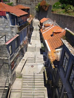 A narrow alleyway lined with old buildings on either side, featuring stone steps descending through the middle. The buildings have rustic elements with tiled orange roofs and stone walls, creating a charming and atmospheric scene. Plants overhang from a stone wall in the background, adding a touch of greenery.