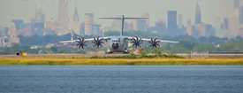 A large military aircraft with four propeller engines is positioned on a runway with an urban skyline featuring various skyscrapers in the background. The foreground contains a body of water and a grassy area bordering the runway, with clear blue skies overhead.
