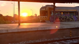A sunset scene with warm orange and yellow hues illuminating a train station platform. A train, featuring graffiti on its side, is parked on the tracks. The sun is low in the sky, casting long shadows and creating a lens flare. In the background, trees and streetlights are silhouetted against the colorful sky.
