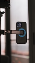 A smartphone is secured on a mount with a blue grip attachment. The setting appears to be indoors, with a window in the background providing soft, diffused lighting.
