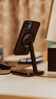 A sleek, foldable phone stand holding a smartphone on a modern wooden desk.