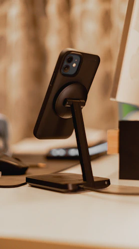 A sleek black mobile phone stand holding a smartphone at a perfect viewing angle on a wooden desk.