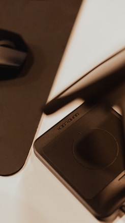 A close-up view of a digital drawing tablet with a stylus resting on it. The design appears sleek and modern with a predominantly black surface, highlighted by the presence of a circular touchpad. The lighting gives it a warm and focused look, with shadows creating a subtle dramatic effect.
