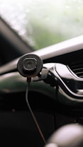 A close-up view of a smartphone holder attached to a car dashboard. The holder is black and cylindrical, and is secured with a cable. The background is softly blurred, showing hints of green, possibly indicating a view through the windshield.