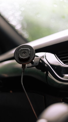 A close-up view of a smartphone holder attached to a car dashboard. The holder is black and cylindrical, and is secured with a cable. The background is softly blurred, showing hints of green, possibly indicating a view through the windshield.