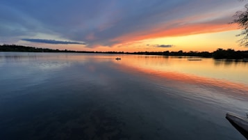 A serene lake scene at sunset with vibrant colors reflecting on the water&rsquo;s surface. The sky features a dramatic gradient from orange to blue, accompanied by scattered clouds. Silhouettes of trees line the horizon, and a small boat is visible on the calm waters.