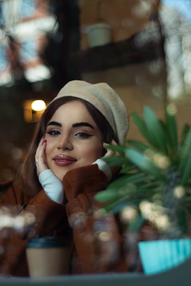 A woman is sitting indoors, resting her face in her hands and smiling softly. She is wearing a beige beret and a brown coat. In the foreground, there is a green plant and a takeaway coffee cup. The background is blurred, suggesting a cozy, relaxed setting.