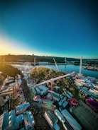 A vibrant amusement park near a body of water with various rides such as a Ferris wheel, roller coasters, and other attractions. The sun is setting, casting a warm glow over the area. Crowds of people move along pathways between colorful tents and stalls, creating a lively atmosphere.