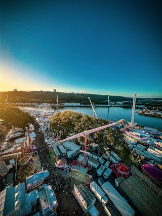 A vibrant amusement park near a body of water with various rides such as a Ferris wheel, roller coasters, and other attractions. The sun is setting, casting a warm glow over the area. Crowds of people move along pathways between colorful tents and stalls, creating a lively atmosphere.