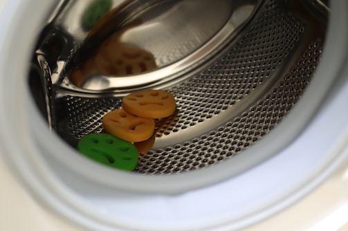 A close-up view of the inside of a washing machine drum with three small, round, smiley face objects, two orange and one green, resting on the metallic surface.