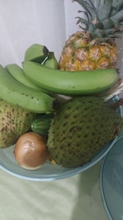 A variety of fruits displayed in a blue wire basket include a ripe pineapple, several green bananas, a soursop, an onion, and a cucumber. The setup is placed against a light background, highlighting the textures and colors of the fruits.