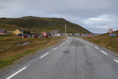 A rural road stretches into the distance with a speed limit sign displaying 70. On either side of the road, grassy fields border a small town with colorful houses, including red and yellow buildings. The scene is set against a backdrop of low, rolling hills under a cloudy sky.