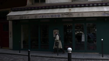 A person walks past a storefront with dark green doors and an old, worn canopy displaying the name 'La Solita Taverna'. The cobblestone street in front adds a historical touch, while the warm glow from inside the store suggests coziness.