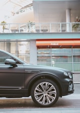 A luxury car is parked in an urban setting, showcasing the front passenger side and wheel design. The background features a modern building with glass panels and an orange awning labeled 'PUBLIC'. Reflections and lighting suggest a sunny day.