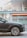 A luxury car is parked in an urban setting, showcasing the front passenger side and wheel design. The background features a modern building with glass panels and an orange awning labeled 'PUBLIC'. Reflections and lighting suggest a sunny day.