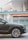 A luxury car is parked in an urban setting, showcasing the front passenger side and wheel design. The background features a modern building with glass panels and an orange awning labeled 'PUBLIC'. Reflections and lighting suggest a sunny day.
