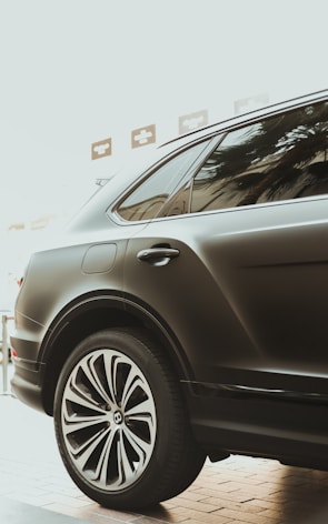 A sleek, black luxury car is parked indoors, showcasing its prominent and stylized alloy wheel. The glossy finish and elegant design reflect modern automotive craftsmanship. The window reflects palm tree silhouettes, adding a touch of sophistication.