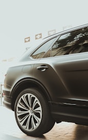 A sleek, black luxury car is parked indoors, showcasing its prominent and stylized alloy wheel. The glossy finish and elegant design reflect modern automotive craftsmanship. The window reflects palm tree silhouettes, adding a touch of sophistication.