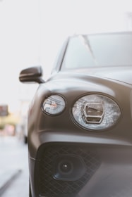 Close-up view of the front left section of a luxury car, focusing on the headlight assembly and side mirror. The car's surface has a matte finish, reflecting some ambient light. The background shows an urban setting with indistinct buildings and street details.